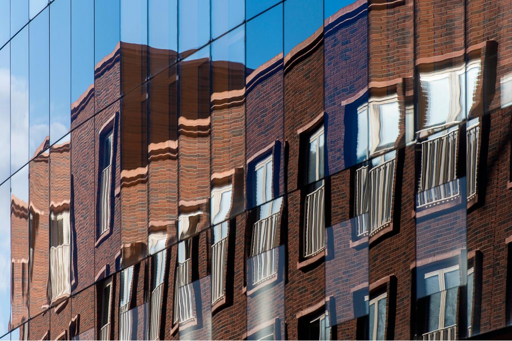 Verzerrte Spiegelung einer Backsteinfassade in moderner Glasfront als Symbol für überlagerte und nicht abgestimmte Nutzungen in innerstädtischen Mischprojekten